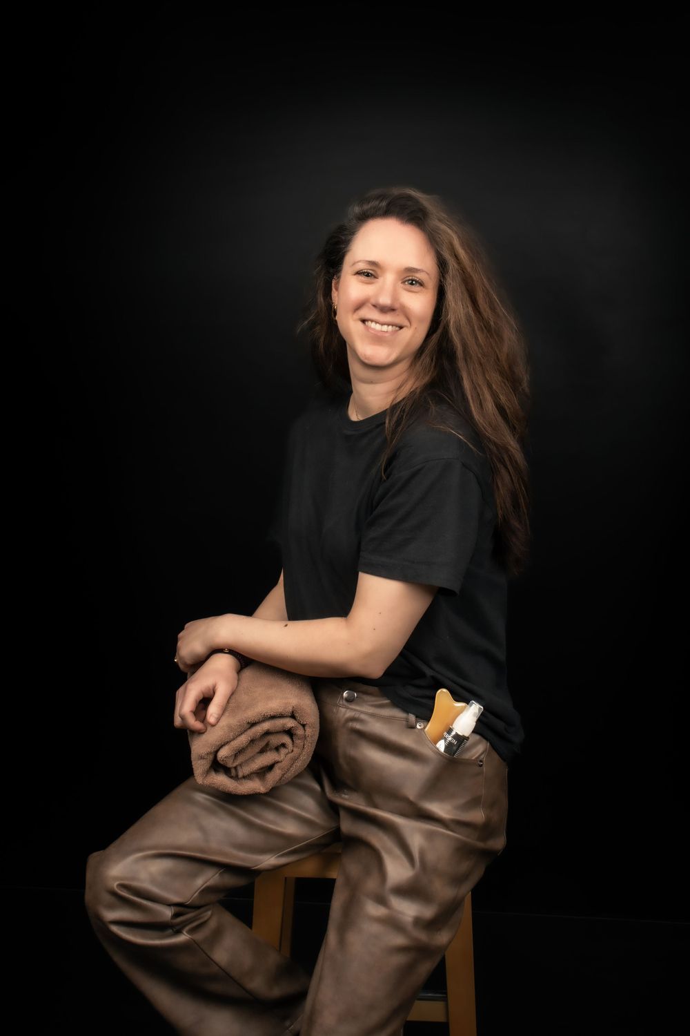 Studio portrait with person in black t-shirt and brown pants sitting casually on wooden stool against dark background.