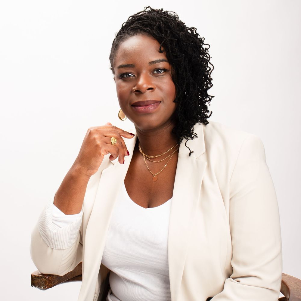 Professional headshot of someone wearing a white blazer and top with natural curly hair against light background.