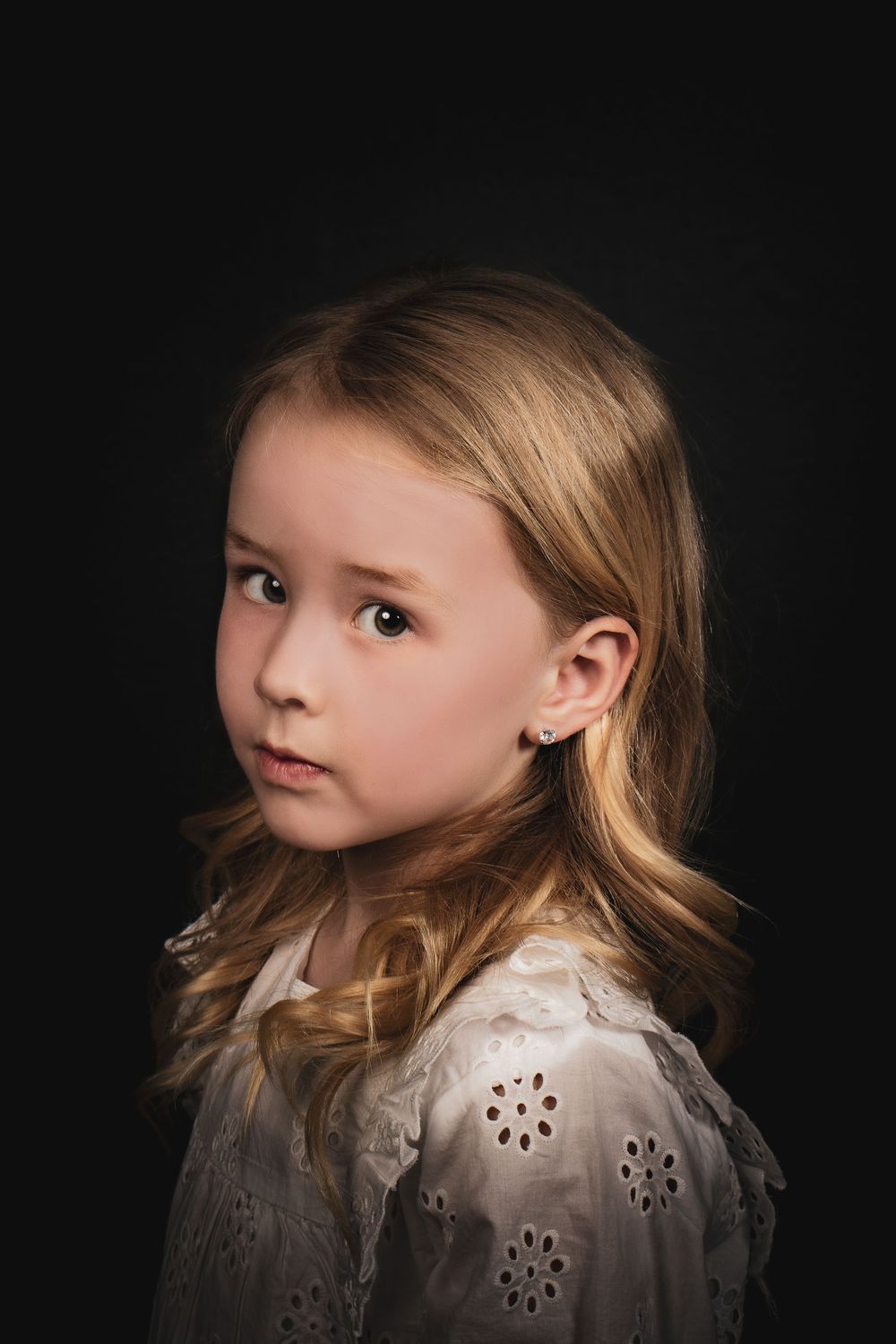 Dramatic portrait of child with blonde hair wearing white floral dress against dark background.