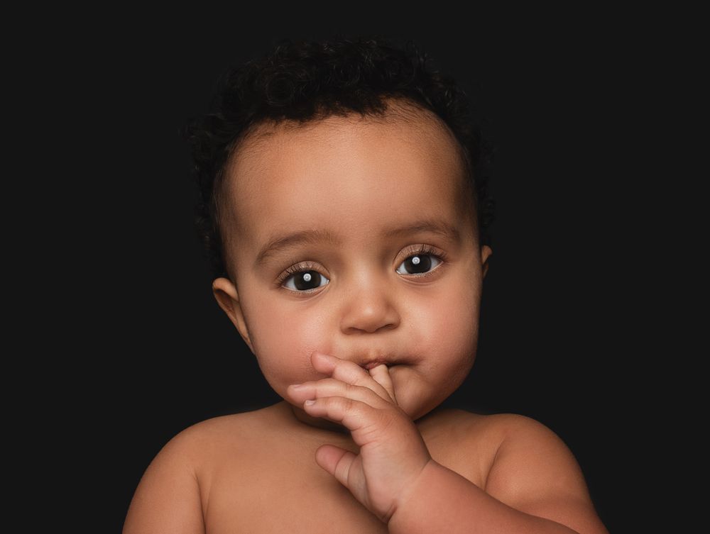 Baby with curly hair and big expressive eyes puts hand near mouth on dark background.