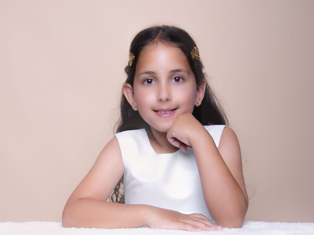 A young student in a white sleeveless top poses gently with hand under chin on light beige background.