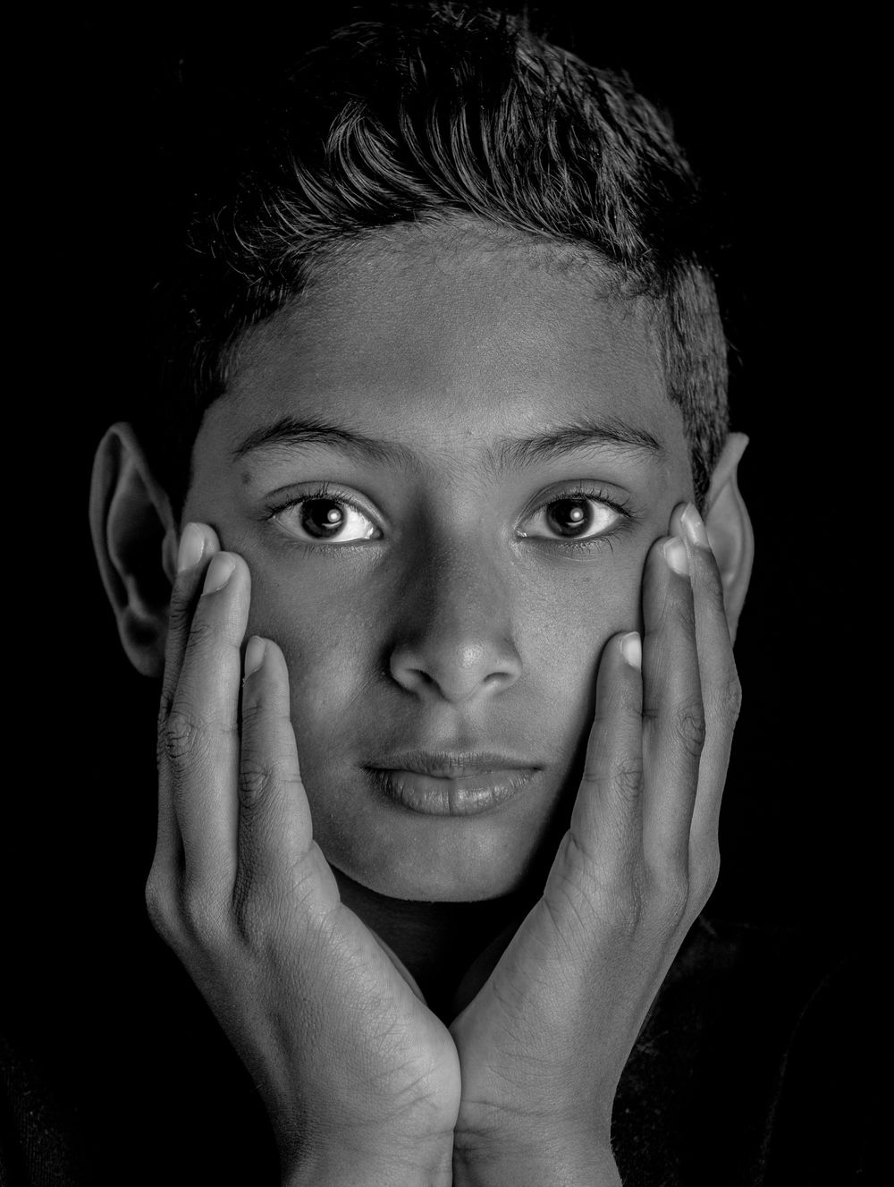 Black and white portrait with hands gently framing a face against dark background in dramatic lighting.