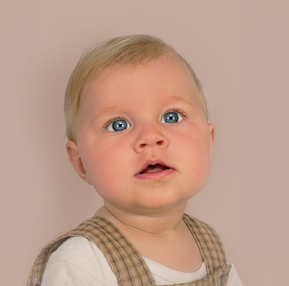 A baby with bright blue eyes wearing a tan vest and white shirt gazes upward with wonder.