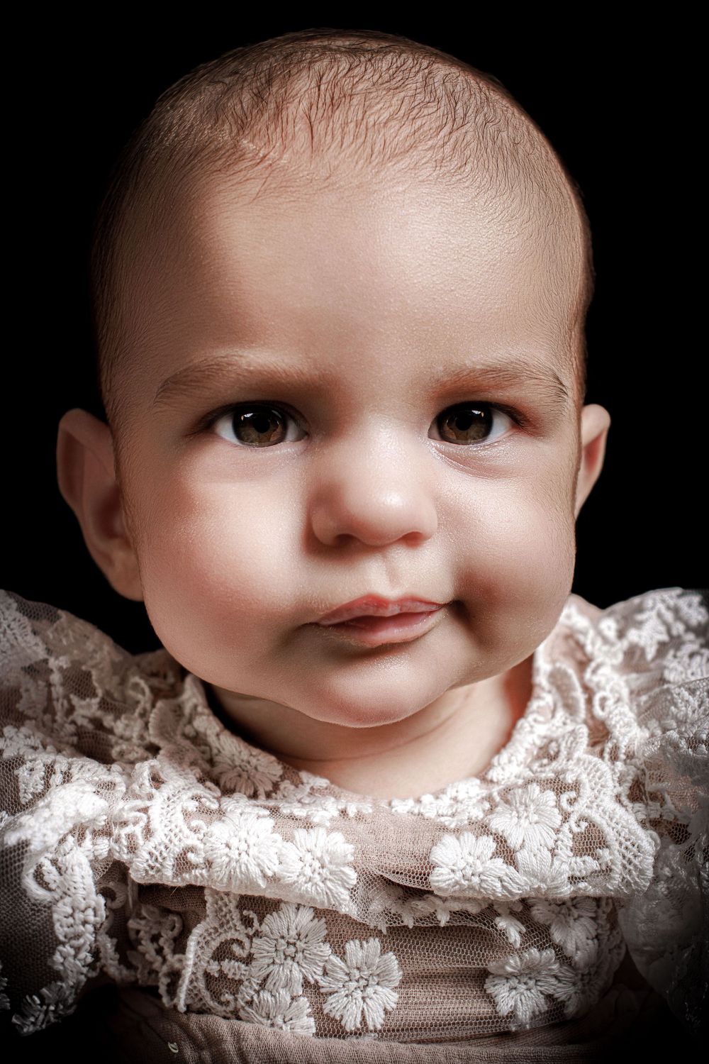 Portrait of a baby wearing delicate white lace against a dark background.