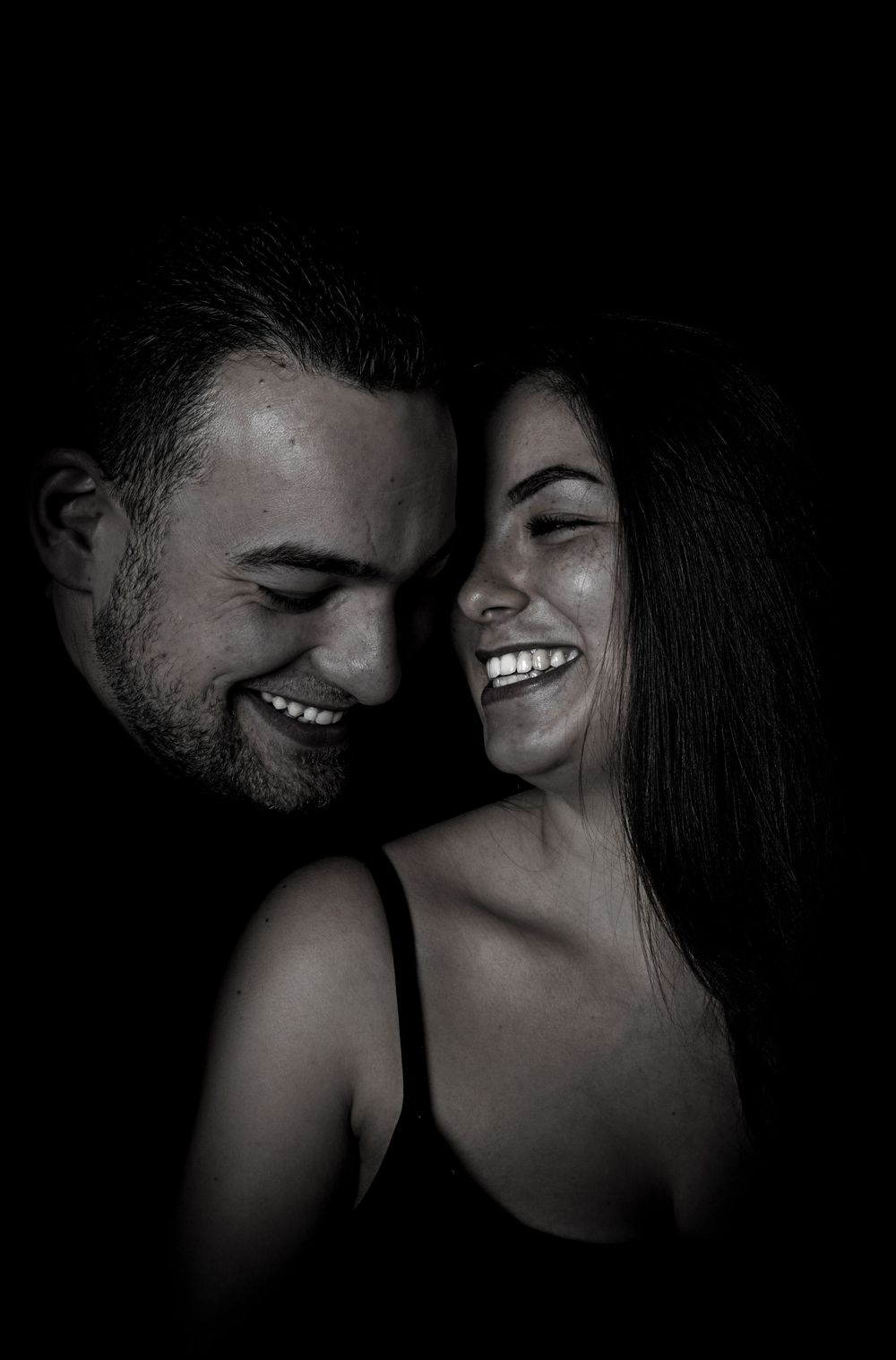 Black and white portrait of a joyful couple laughing together in intimate moment against dark background.