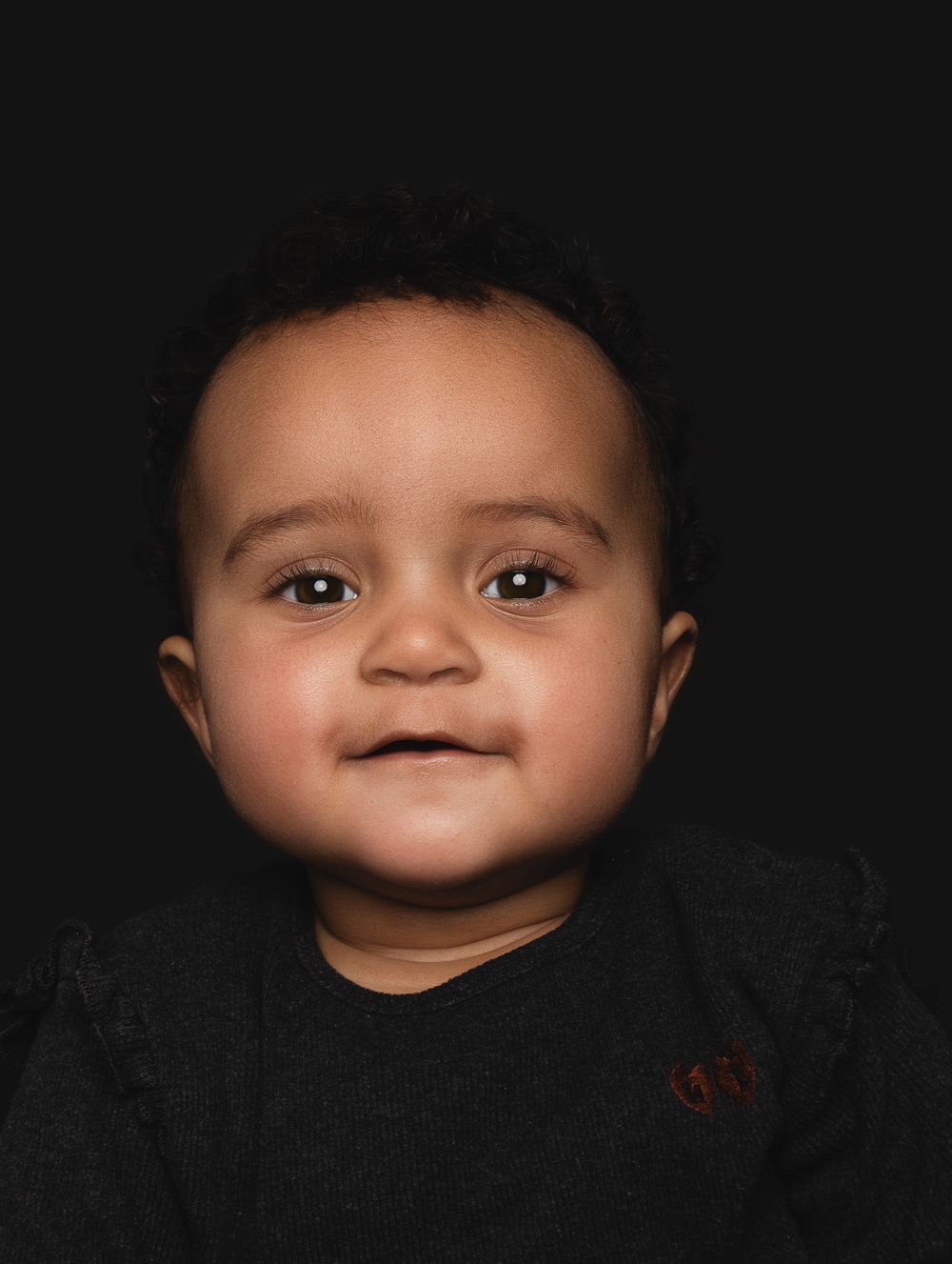 An adorable baby wearing a dark shirt smiles warmly against a simple dark background in a portrait-style photo.