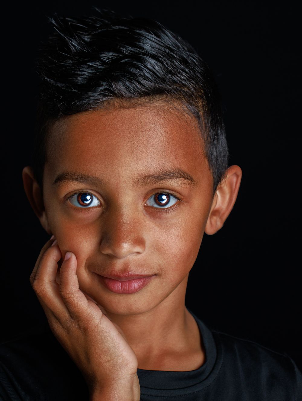 Portrait shot against dark background highlighting striking blue eyes and gentle expression while resting chin on hand.