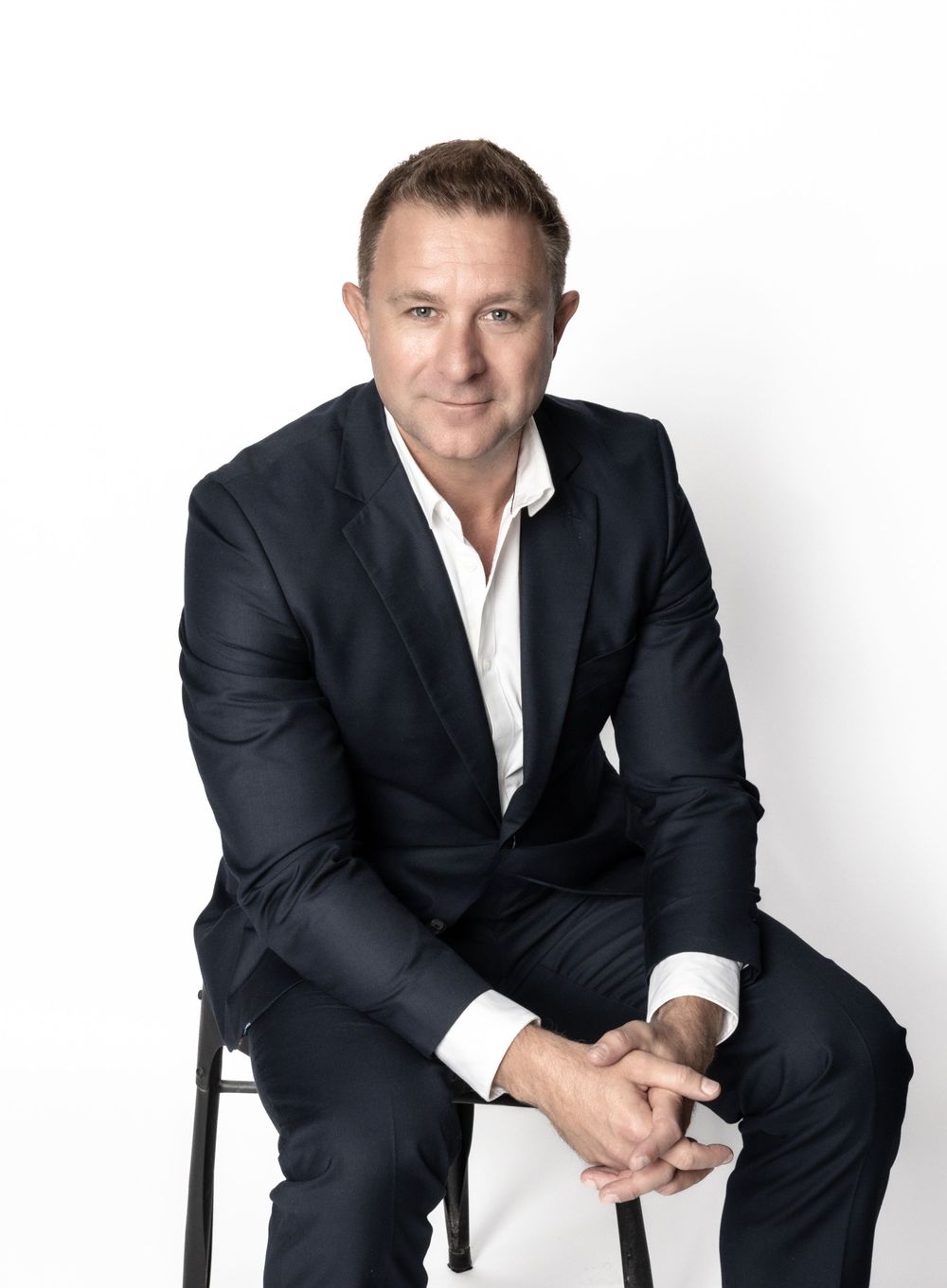 Professional portrait of someone in a black business suit sitting sideways on a chair against a white background.