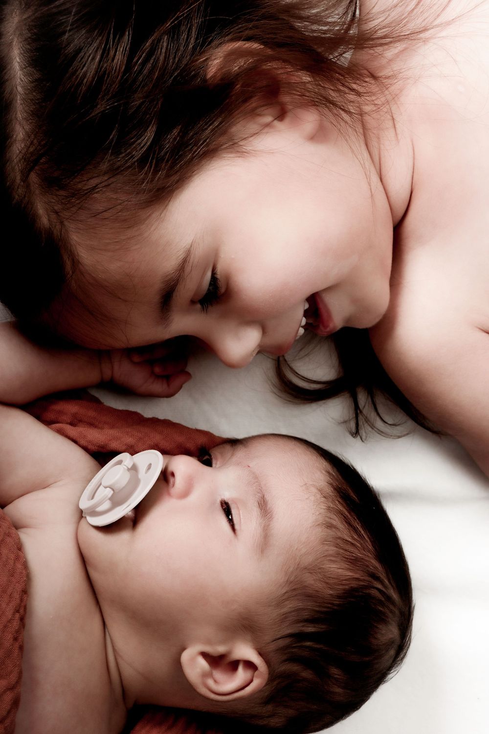 A gentle moment between a baby with a pacifier and an older sibling lying together on a white surface.