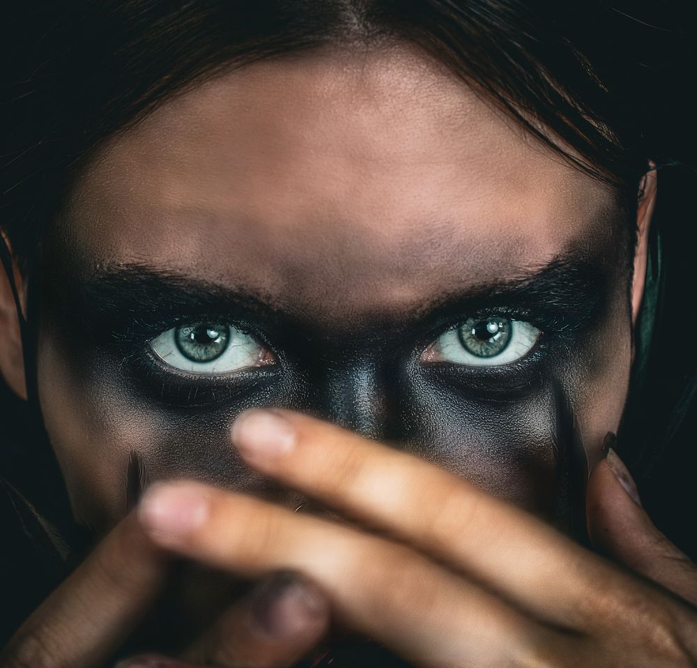 Striking artistic portrait with dramatic black face paint highlighting piercing blue eyes and a hand partially covering the face.