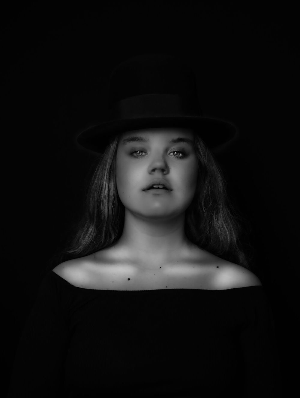 Black and white artistic portrait with dramatic lighting featuring a subject wearing a dark hat against a black background.