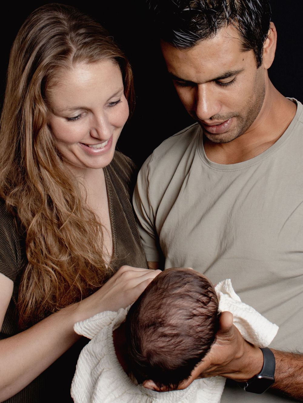 Loving parents smile while holding and gently drying their newborn baby in a dark-toned studio portrait.