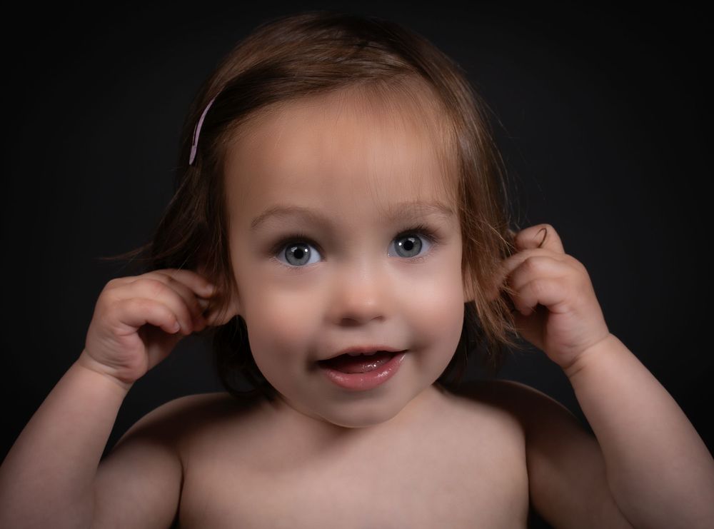 Young child pulls both ears while smiling against dark background in dramatic studio portrait.