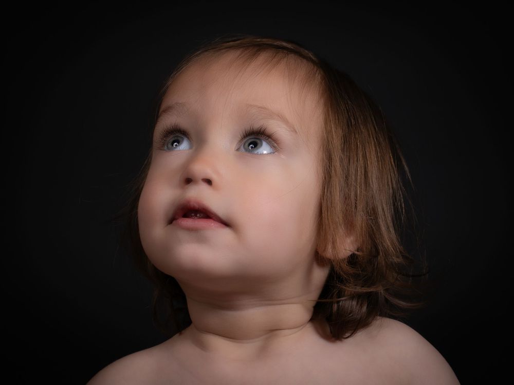 A young child gazing upward against a dark background with soft lighting illuminating their face and shoulders.