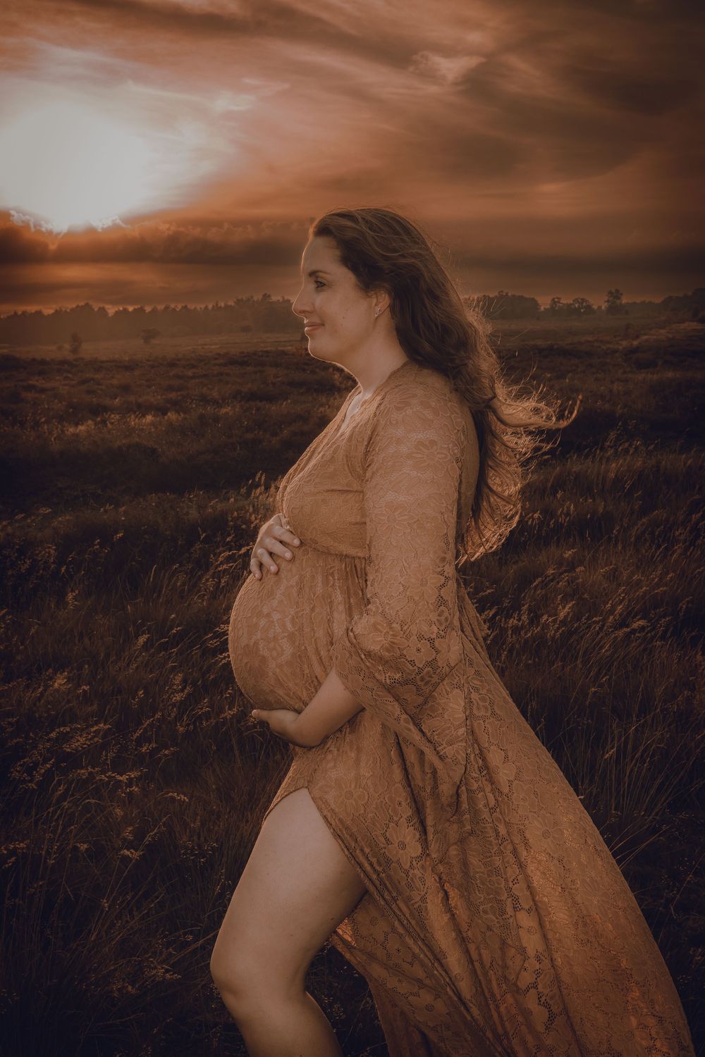 Silhouetted maternity photo in golden sunset light showing pregnant figure in flowing dress standing in grassy field.