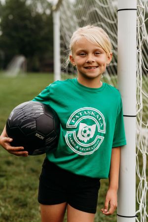 Young soccer player in green Alaska club shirt poses with black ball by goal post during practice on grass field.