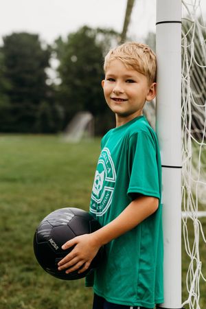Young player in green jersey holds black soccer ball while leaning against goal post on grassy field.
