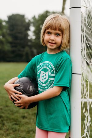 Young player in green soccer team shirt holds black soccer ball while standing by goal post on grass field.