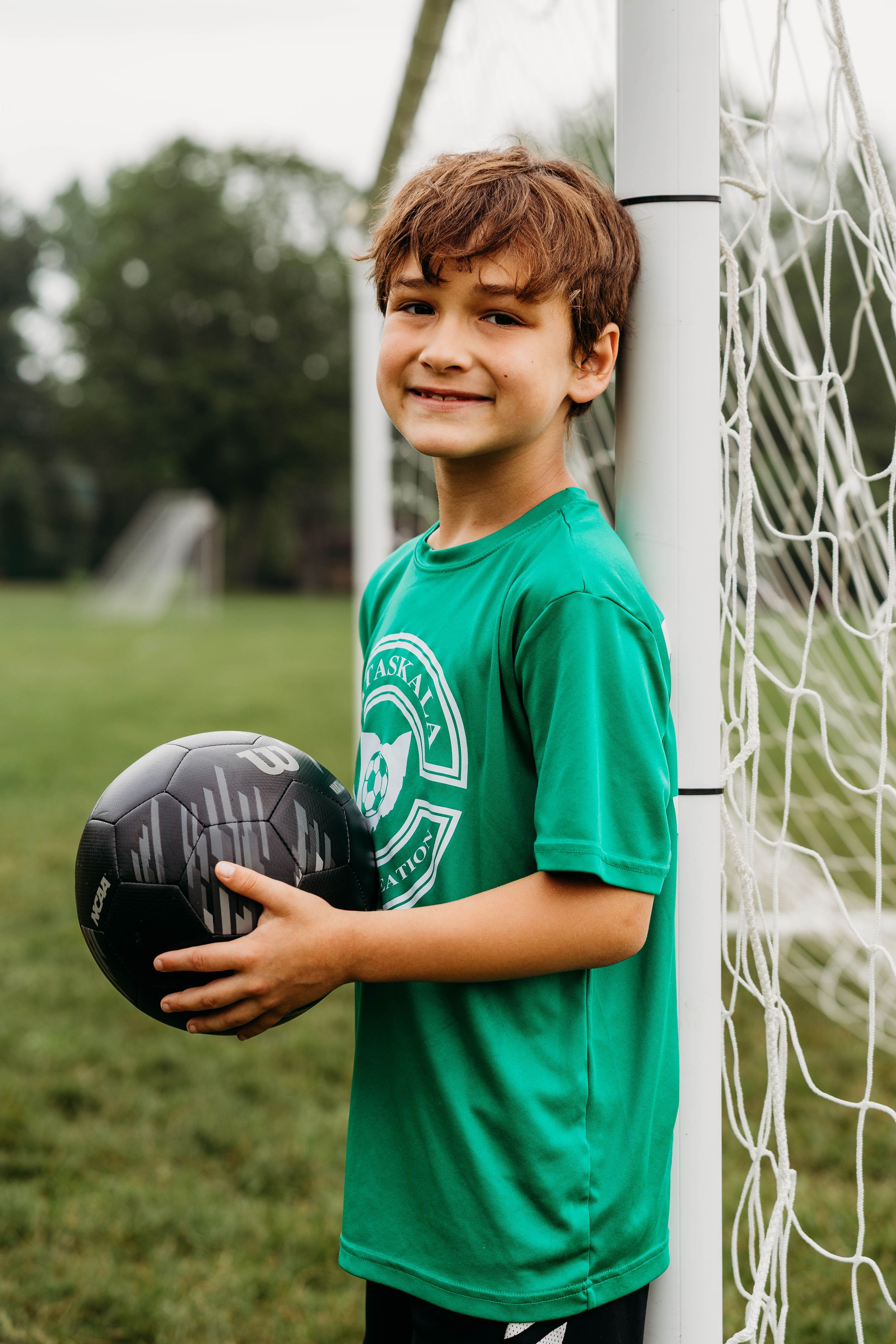 Young player in green jersey holds a black soccer ball while standing next to a goal post on a grassy field.