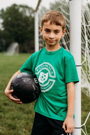 A young soccer player stands by a goal post wearing a green t-shirt with a club logo and holding a black soccer ball.