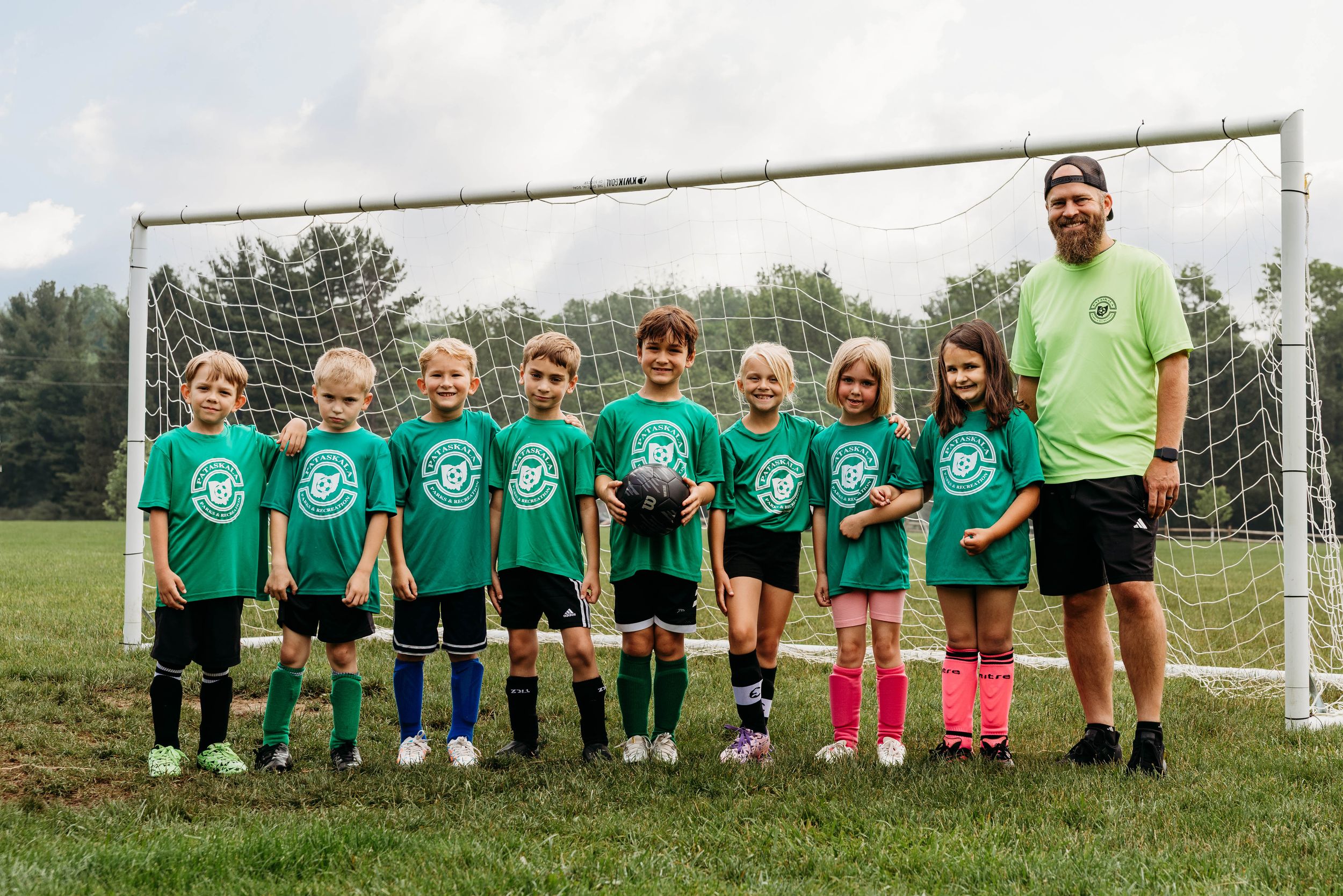 Youth soccer team in green jerseys lines up with their coach in front of a goal on a grassy field.