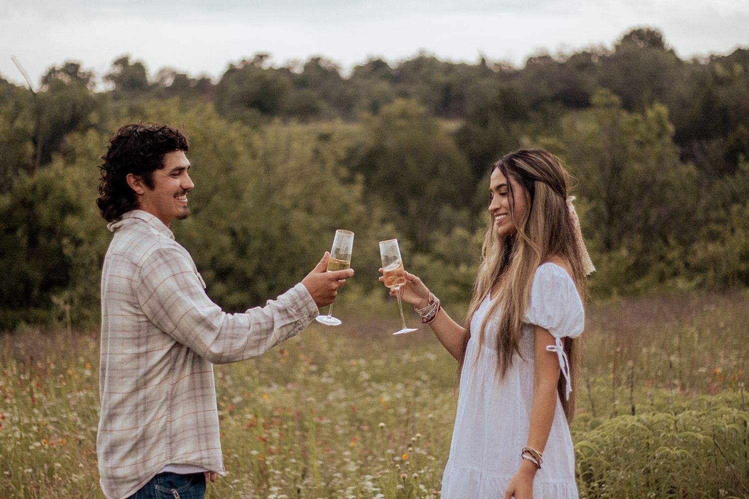 Two people toasting with wine glasses in a golden field during sunset with trees in the background.