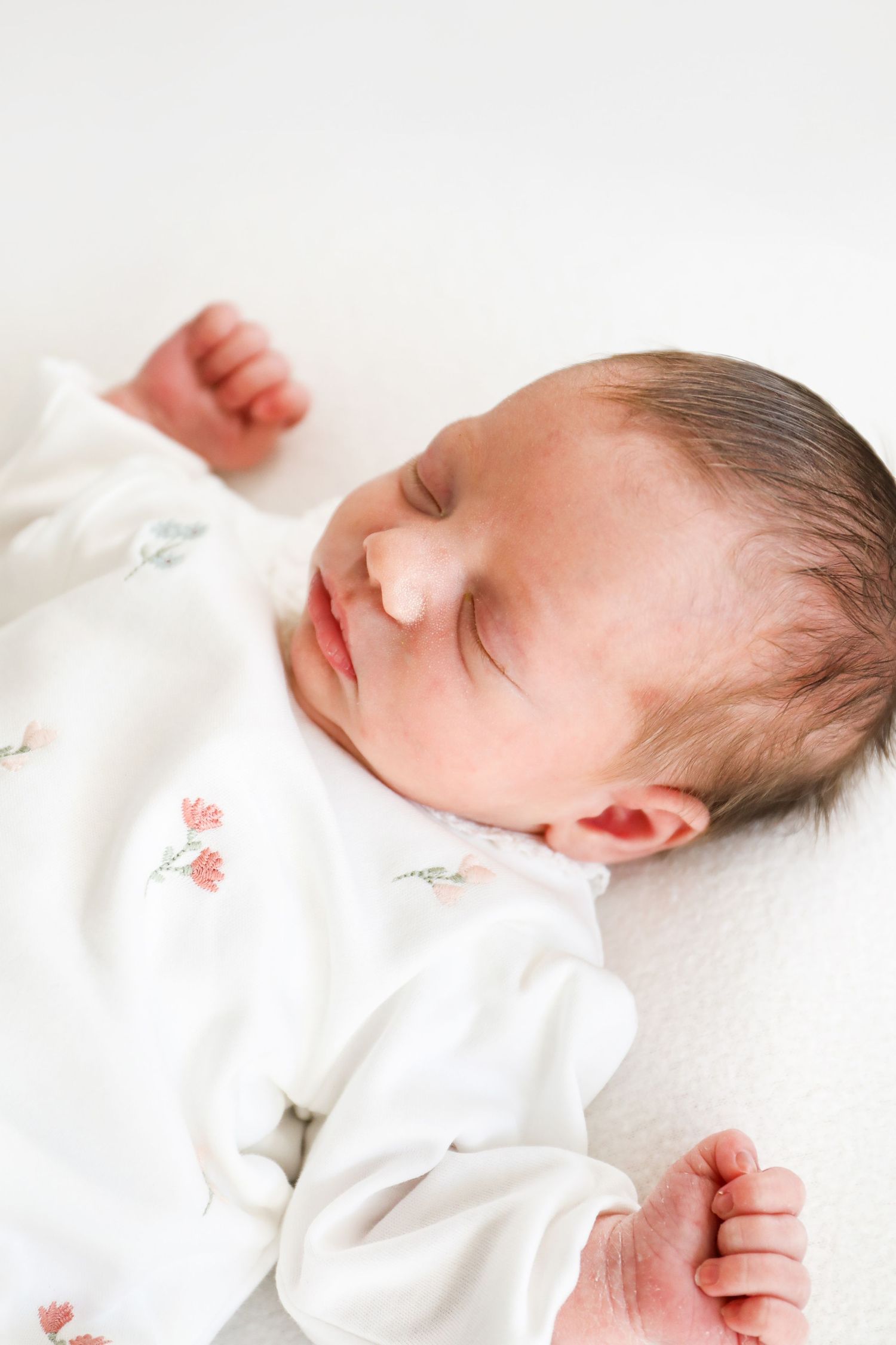 Peaceful sleeping infant wearing white sleepwear with floral design rests on light background during Newborn shoot in Nottingham