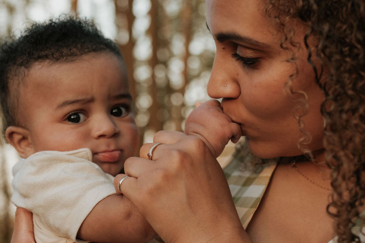 A mother tenderly kisses her baby's hand while the infant gazes curiously at the camera outdoors.