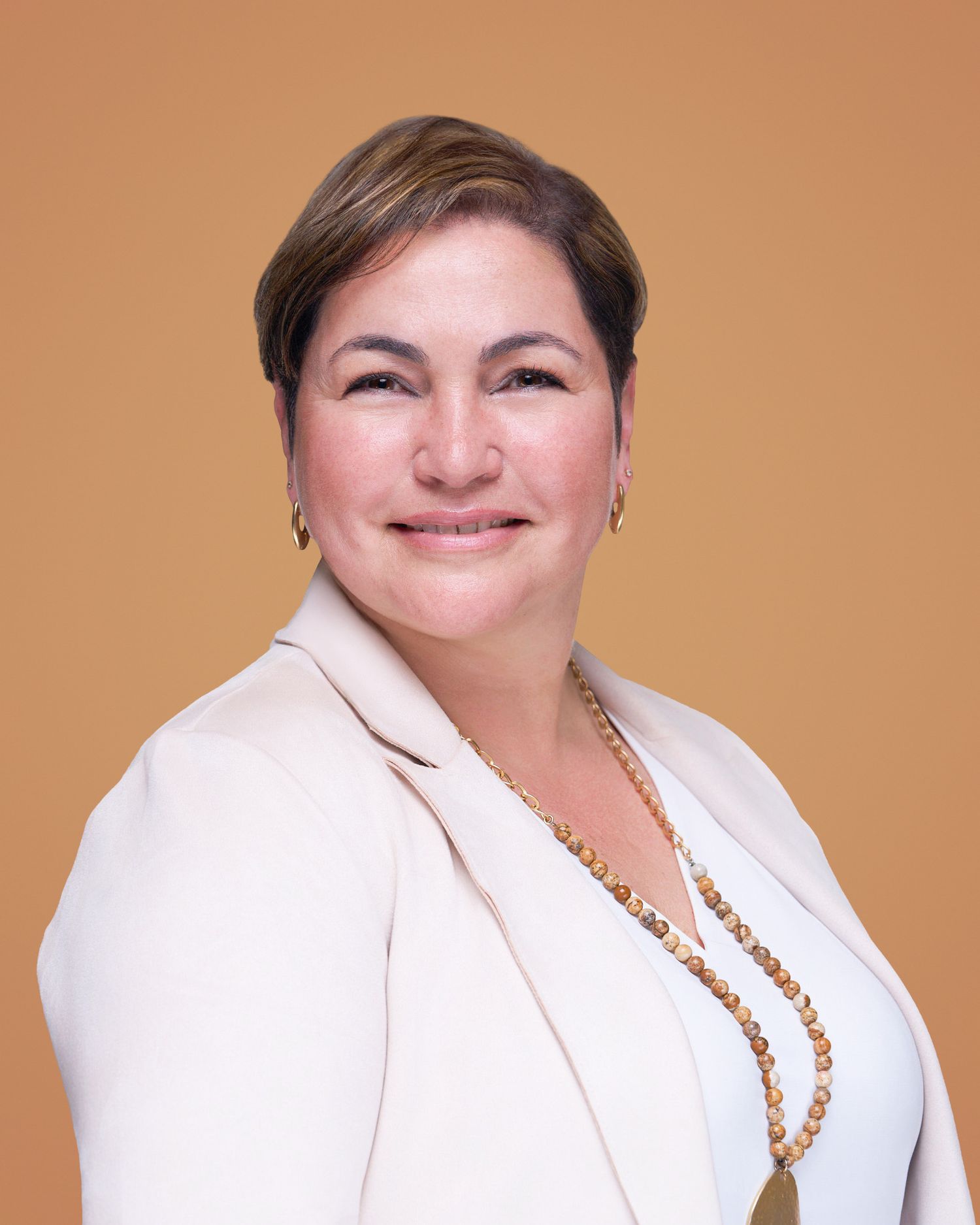 Professional portrait in cream blazer with pearl necklace against orange background.