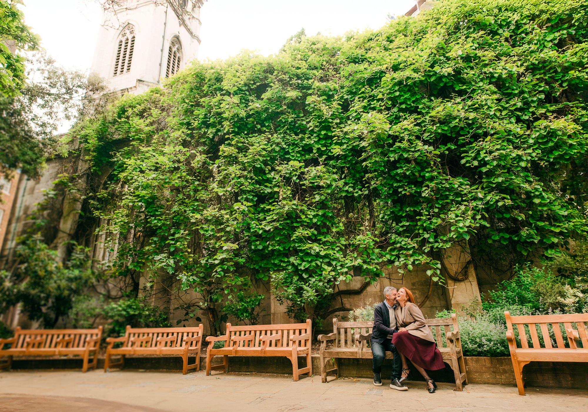 Wooden benches line a peaceful garden path surrounded by lush green foliage and trees at an old church or cathedral.