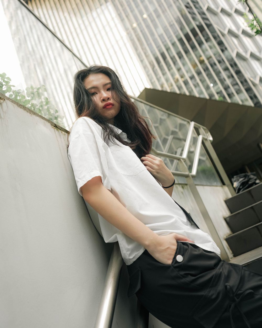 Fashion portrait of a stylish person in a white t-shirt and black pants posing casually on stairs with architectural details.