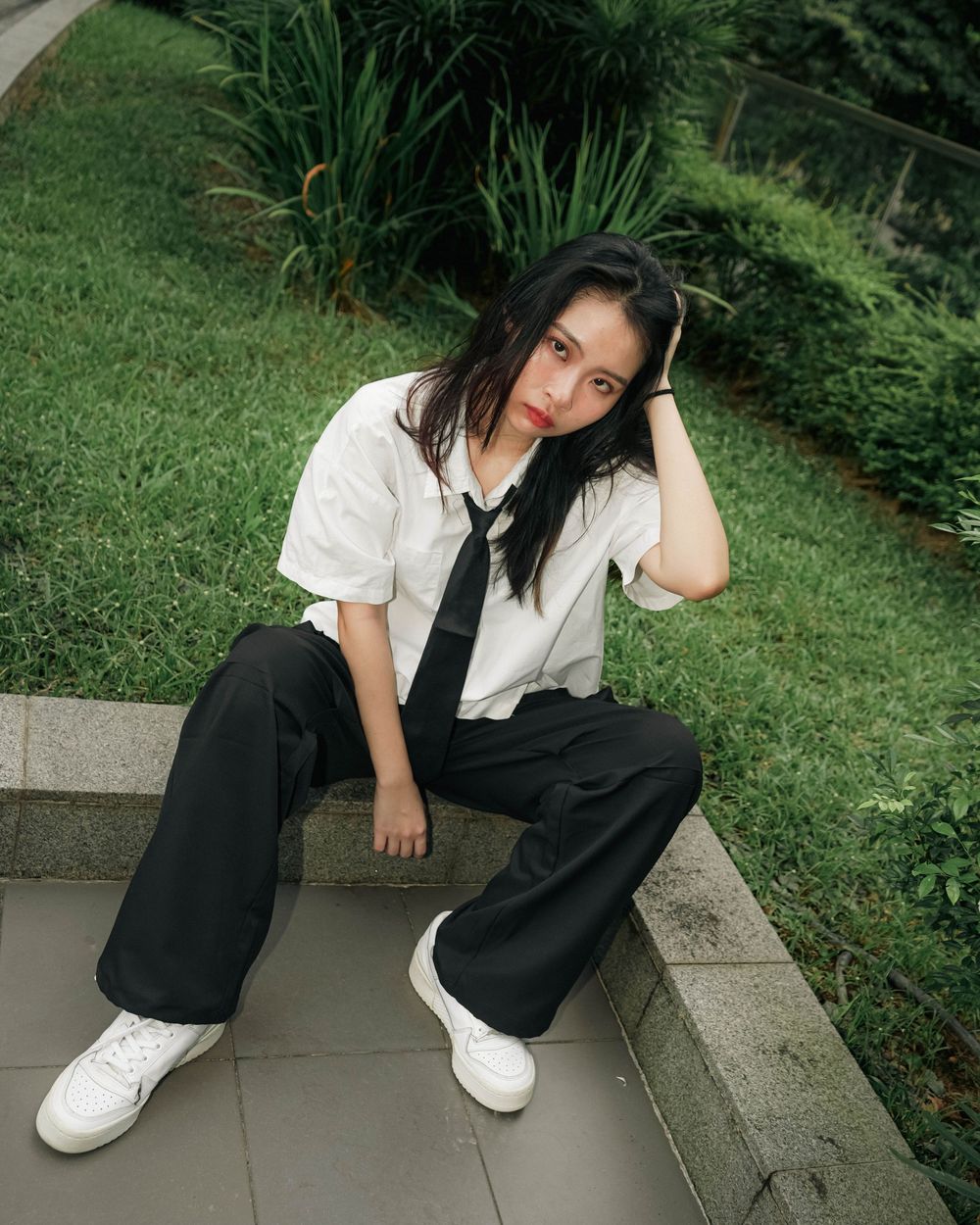 Someone in a white button-down shirt and black pants sits casually on concrete steps in a garden setting.