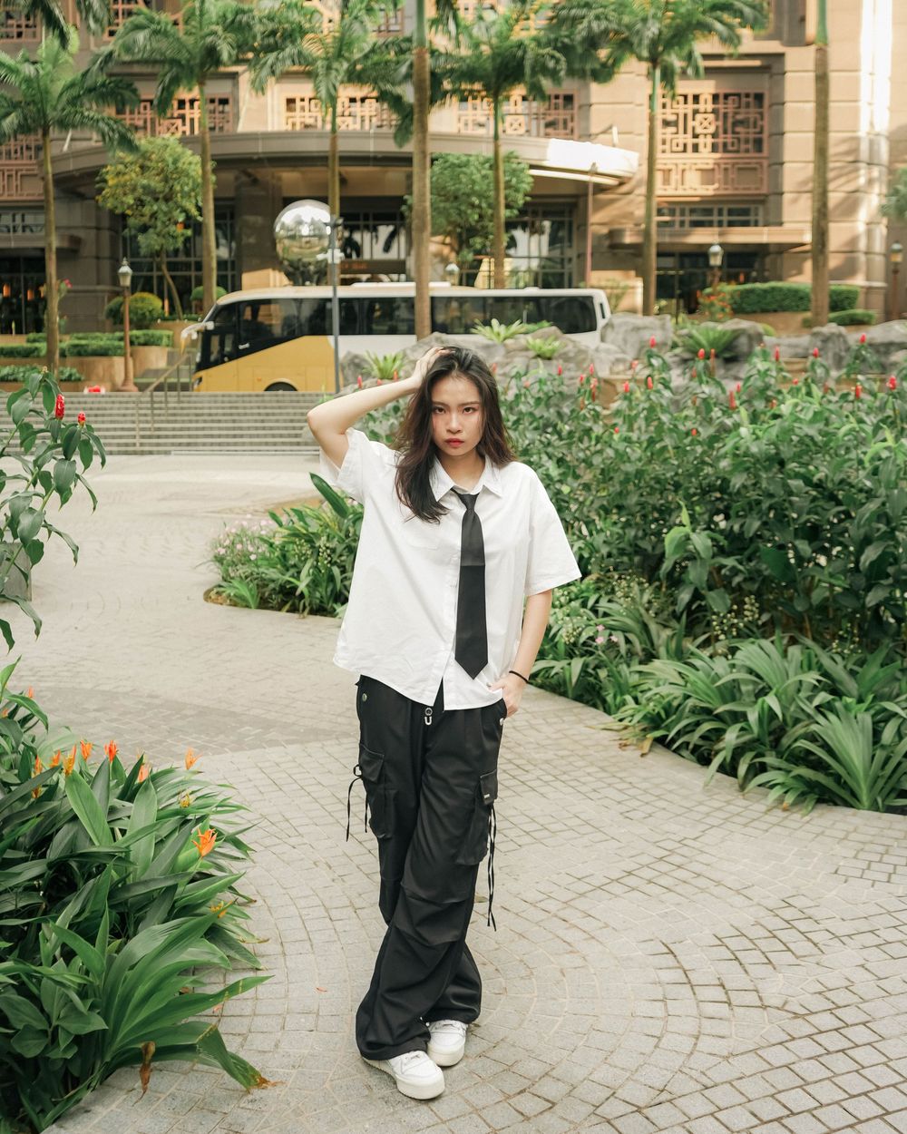 A person in a black and white outfit poses on a curved brick path surrounded by lush greenery and architectural details.