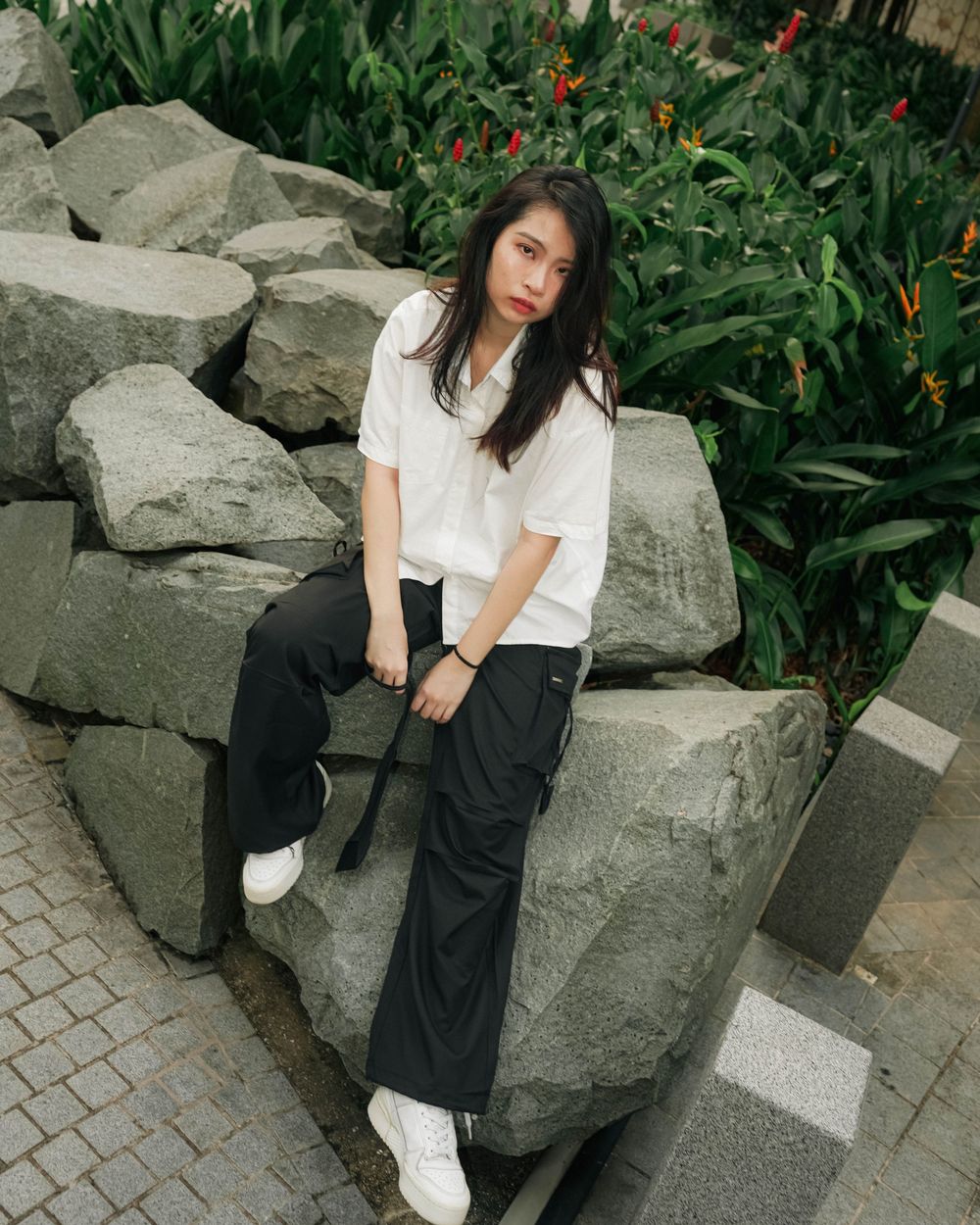 A person in a casual white shirt and dark pants sits on large rocks next to greenery with red flowers.