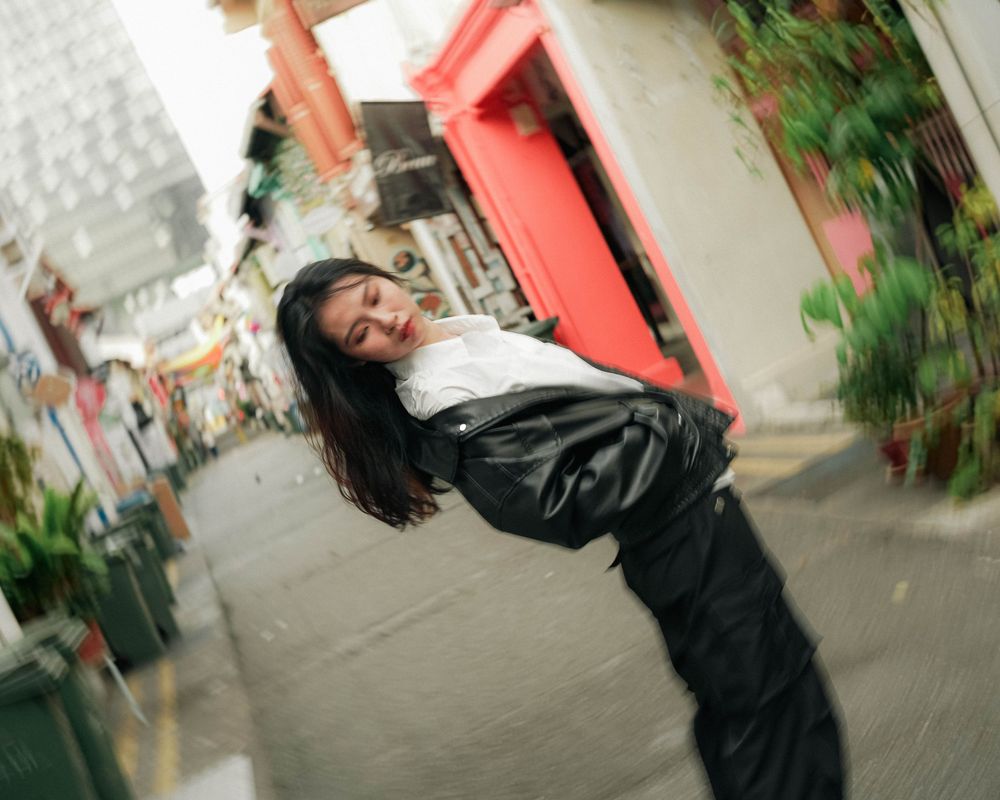 Someone wearing a black jacket and pants poses playfully on a city street with motion blur effects and greenery visible.