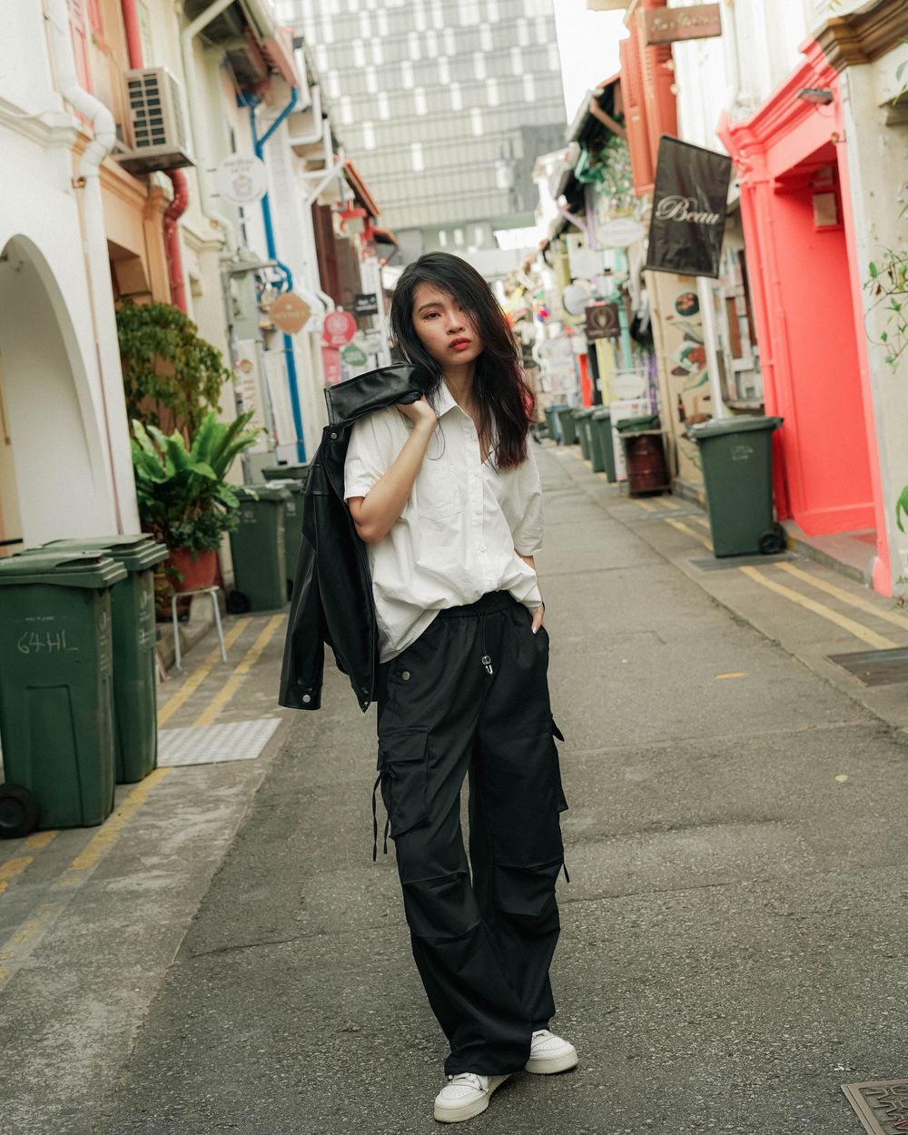Person wearing black jacket and cargo pants poses on charming alley with colorful shophouses and green plants in Singapore.