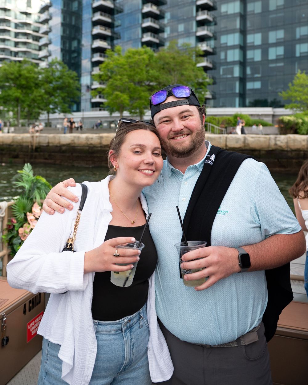 A couple enjoying drinks together on a waterfront deck with city apartment buildings in the background.