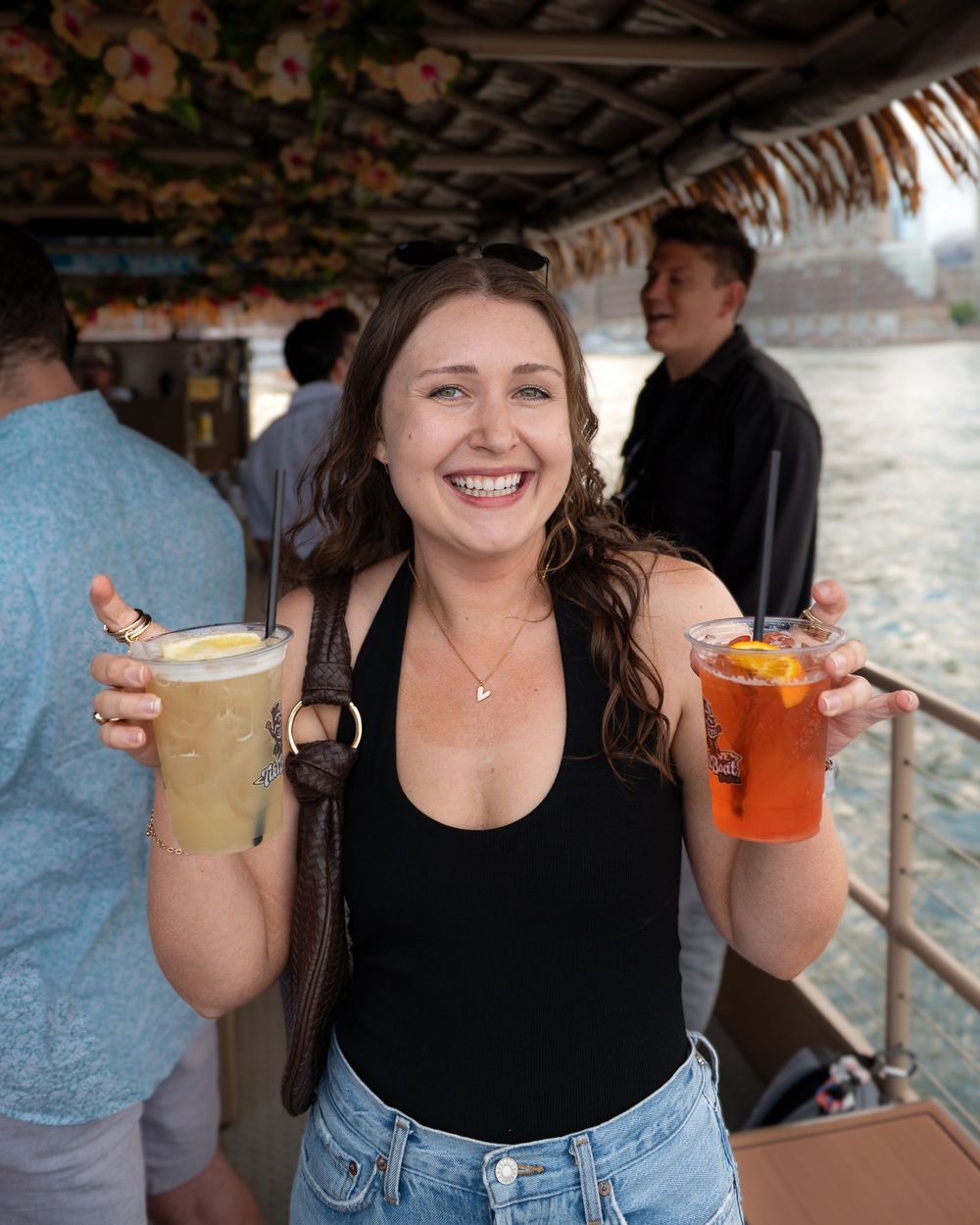 Someone in a black tank top and jeans holds colorful cocktails at a tiki bar with other guests visible in background.