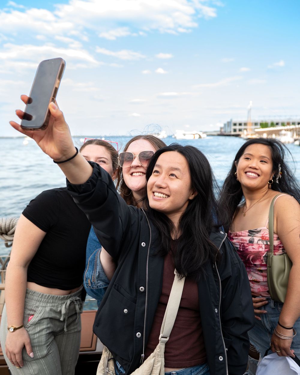 A group takes a selfie together by the waterfront on a sunny day with boats and buildings visible in the background.