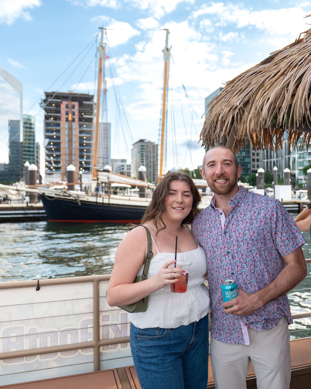Couple enjoying drinks at a waterfront tiki bar with a tall ship and city buildings in the background.