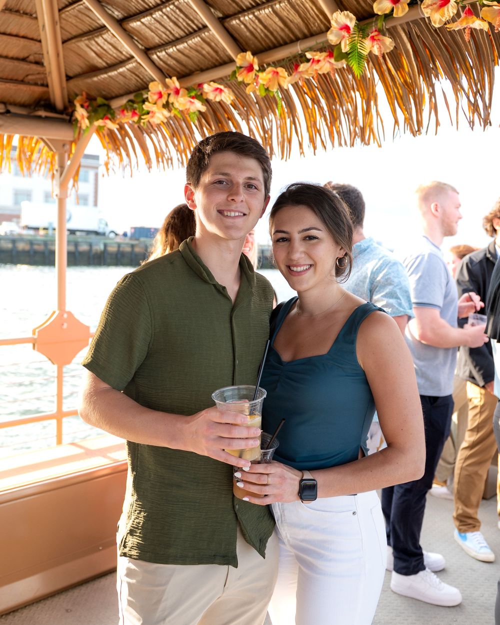 A couple enjoying drinks under a tiki-style thatched roof bar decorated with tropical flowers and foliage.