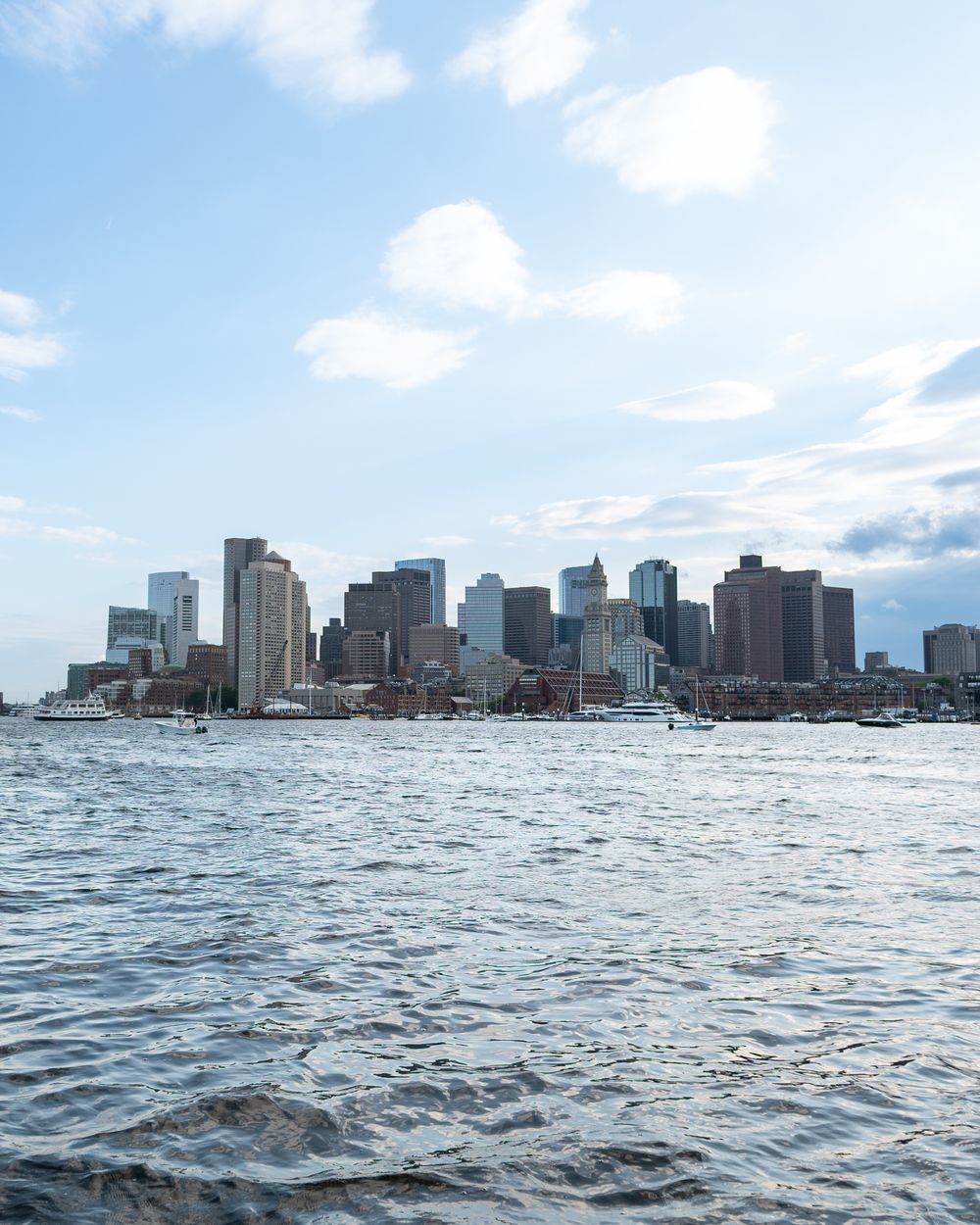Boston skyline towers over the harbor waters on a sunny day with scattered clouds.