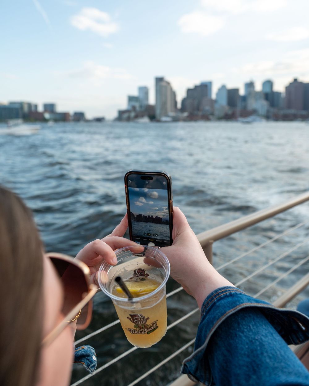 Hands hold phone and drink while enjoying Boston harbor views with city skyline in background on sunny day.