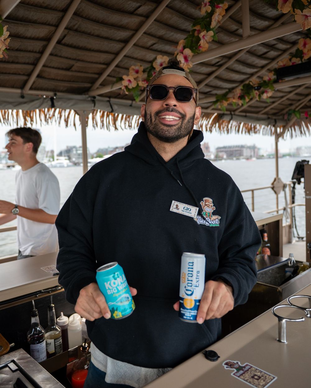 Bartender in black hoodie holds beverage cans while working at a covered tiki bar.