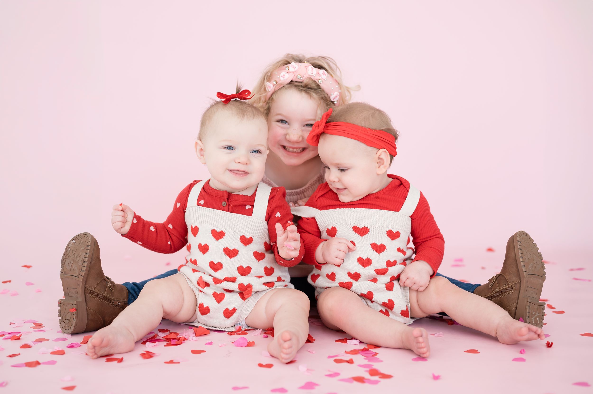 Children in heart-patterned Valentine's Day outfits pose playfully on pink background.