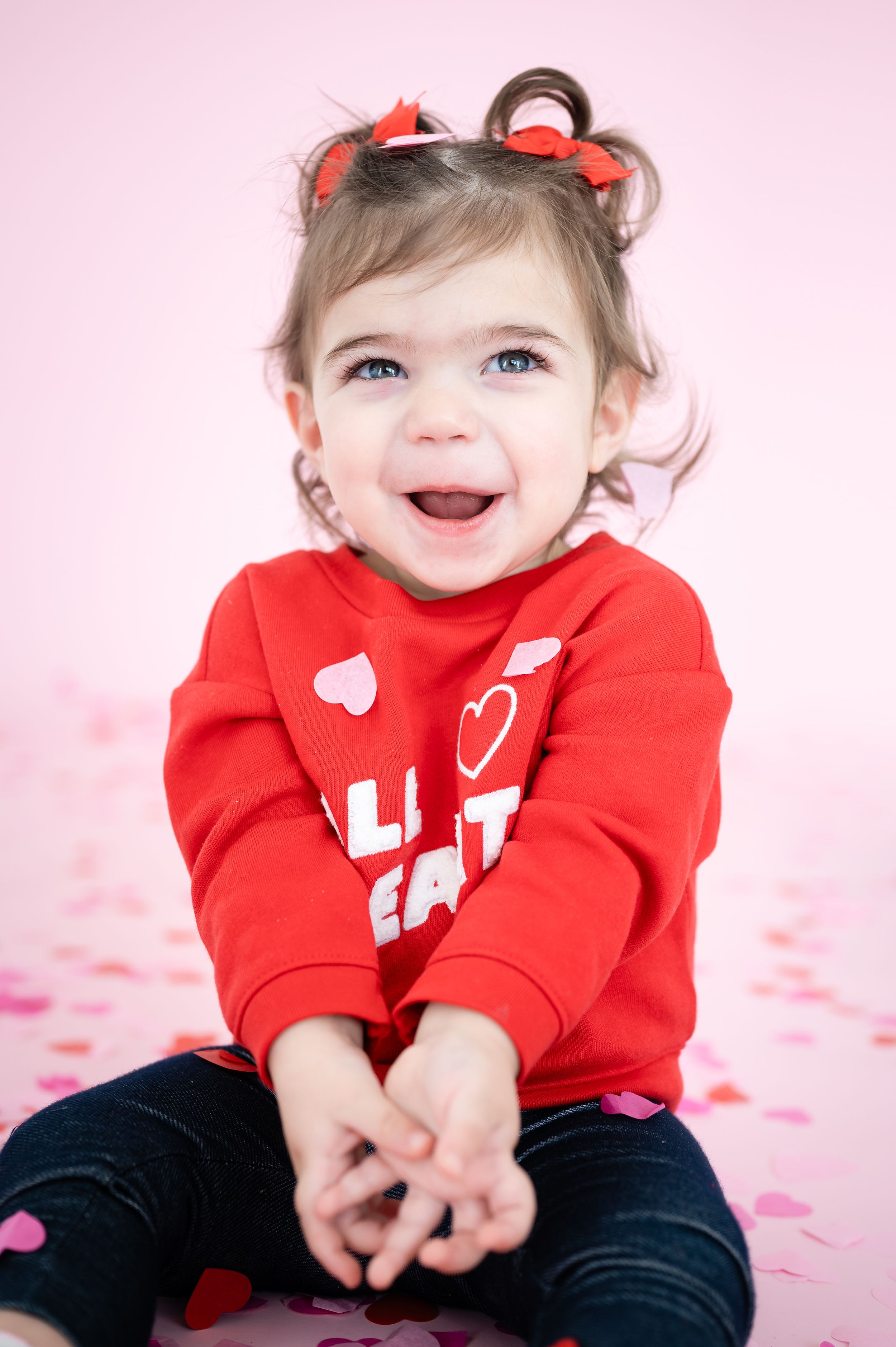 Child in red heart sweater smiles while sitting on pink background with scattered hearts.