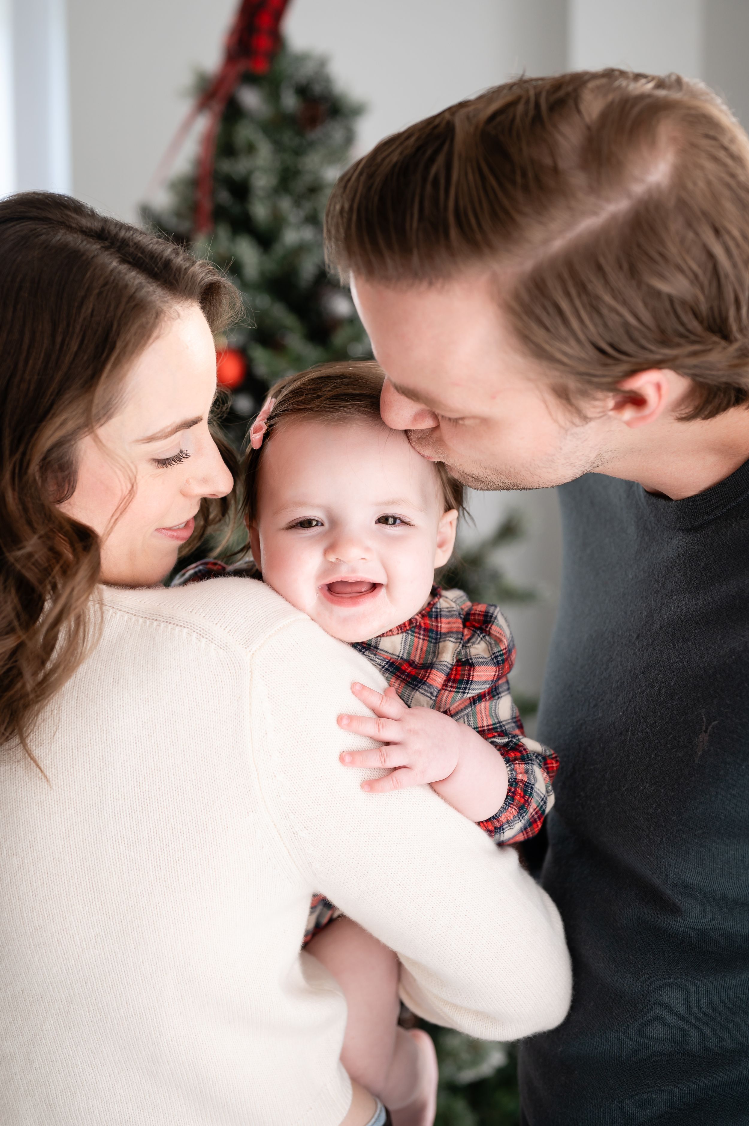A joyful family moment captured indoors with a smiling baby in a plaid outfit during the holiday season.