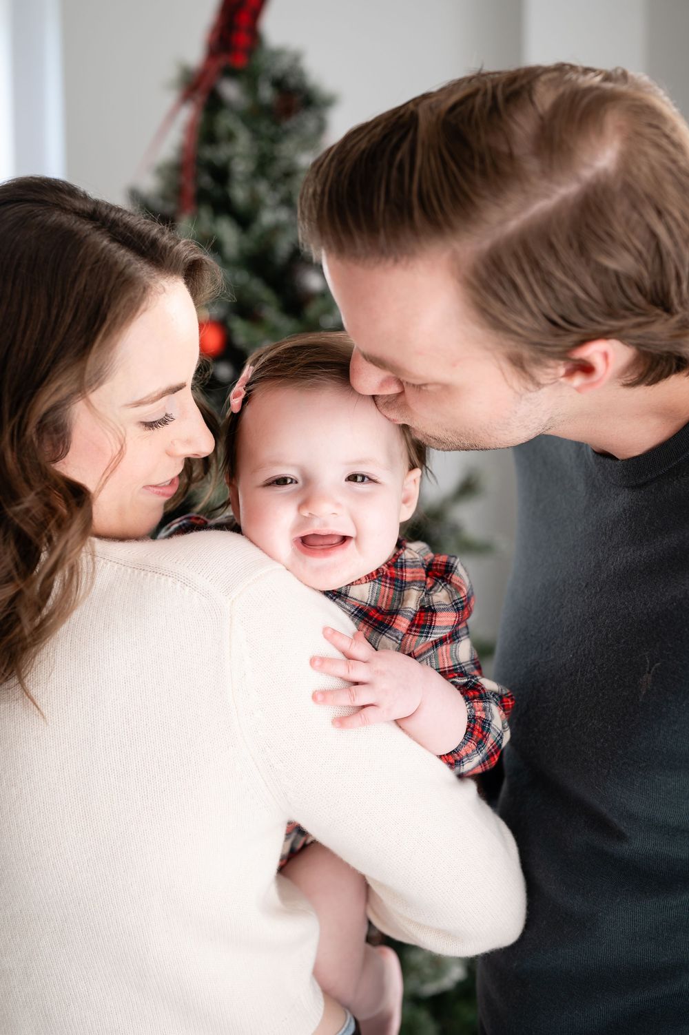 A joyful family moment captured indoors with a smiling baby in a plaid outfit during the holiday season.