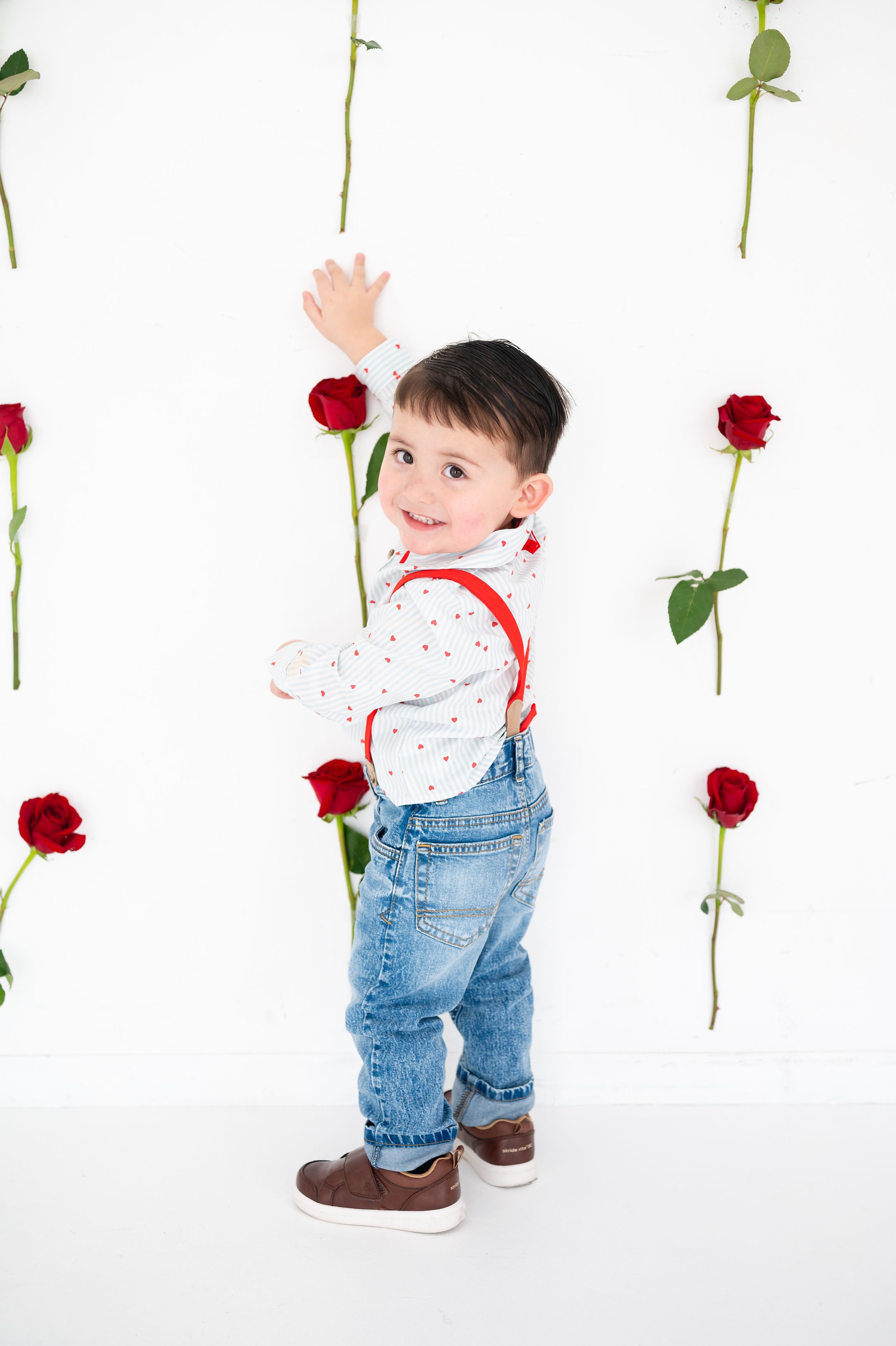 Small child in jeans and suspenders plays among hanging red roses against white background.