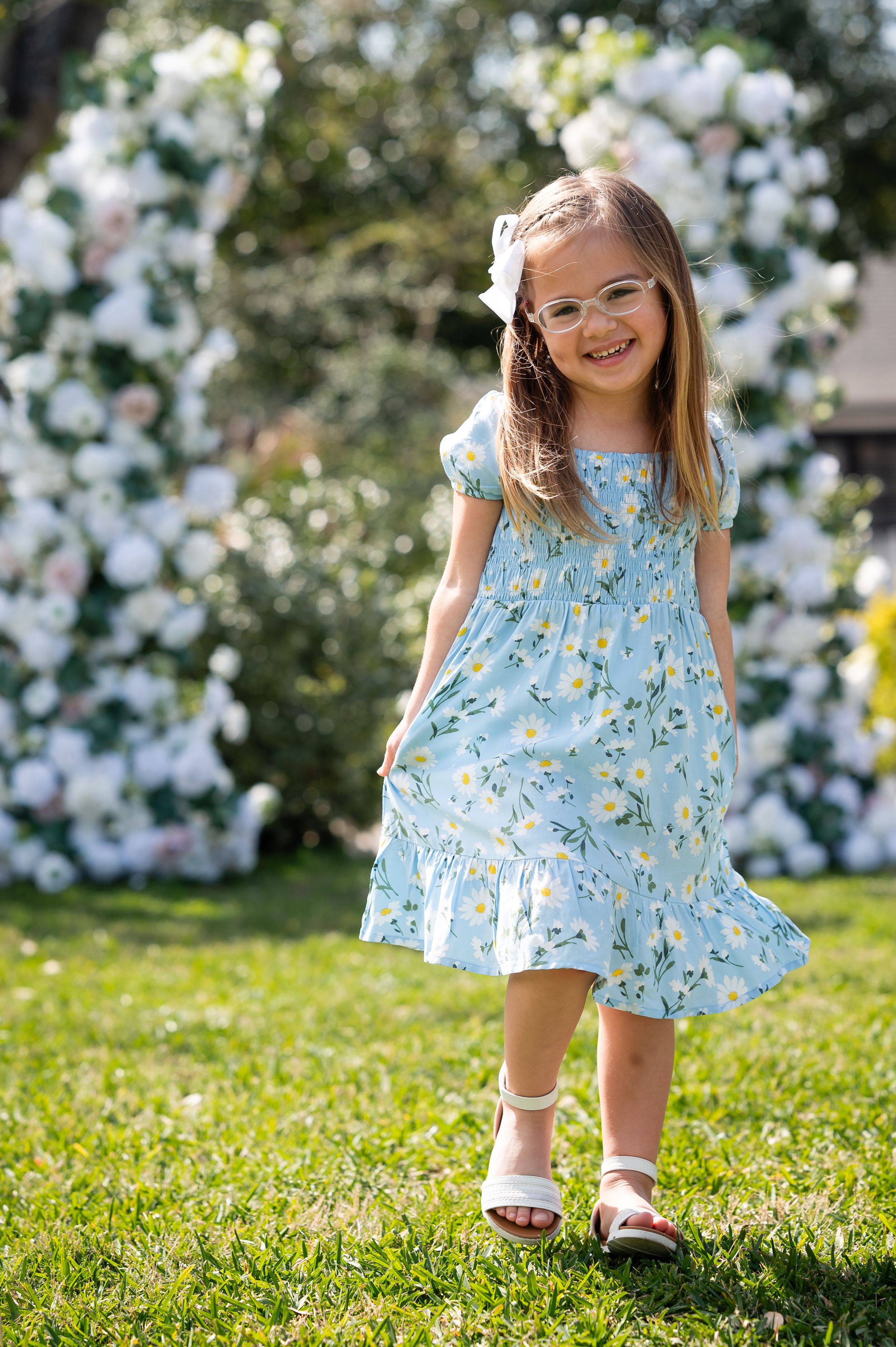 Young child in a blue floral dress twirls playfully on green grass with white flowers in the background.